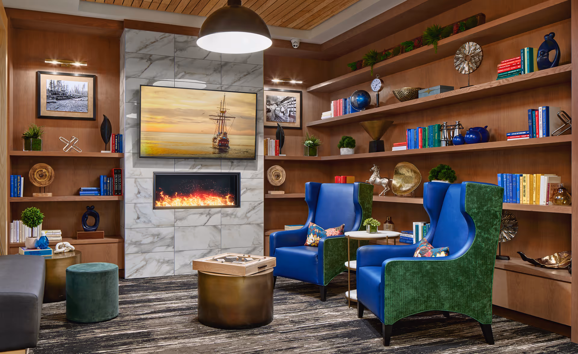A cozy senior living common area featuring two blue and green armchairs with floral pillows, a modern electric fireplace set in a marble wall, a wall-mounted TV displaying a ship at sunset, wooden bookshelves filled with books and decorative items, a small round table, and a patterned carpet.
