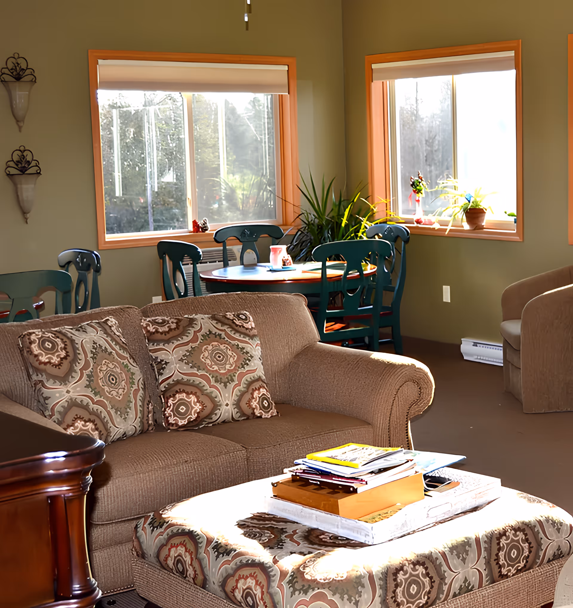 A cozy living room area with a brown sofa adorned with patterned cushions, a matching ottoman with books and magazines on top, and a round wooden dining table surrounded by green chairs near two large windows with beige blinds. There are potted plants on the windowsill and a beige armchair in the corner.