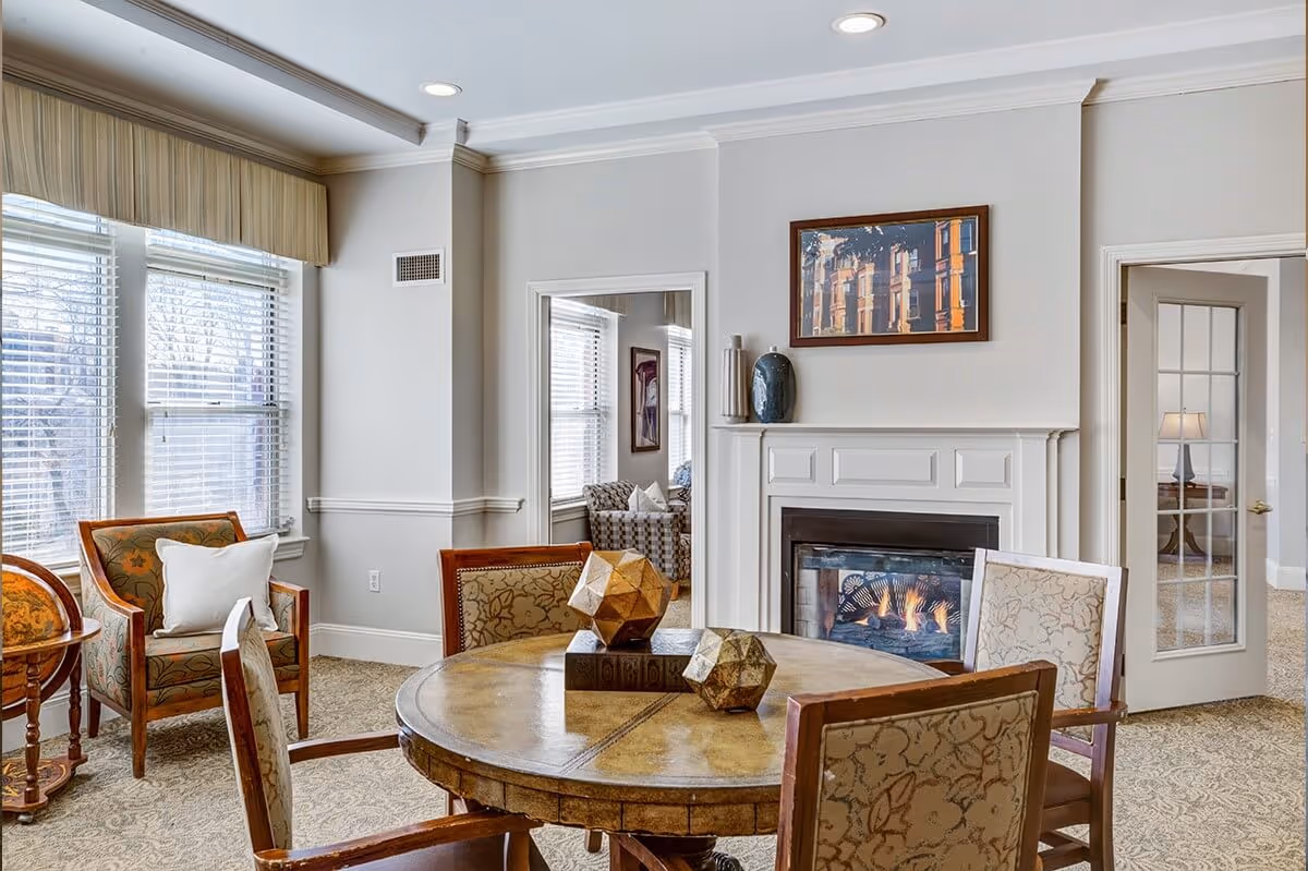 A cozy living room area with a round wooden table surrounded by four upholstered chairs. The room features a lit fireplace with a decorative mantle, a framed picture above it, and large windows with blinds letting in natural light. There is a small globe on a stand near one of the windows and an adjacent room visible through a doorway with glass-paneled double doors.