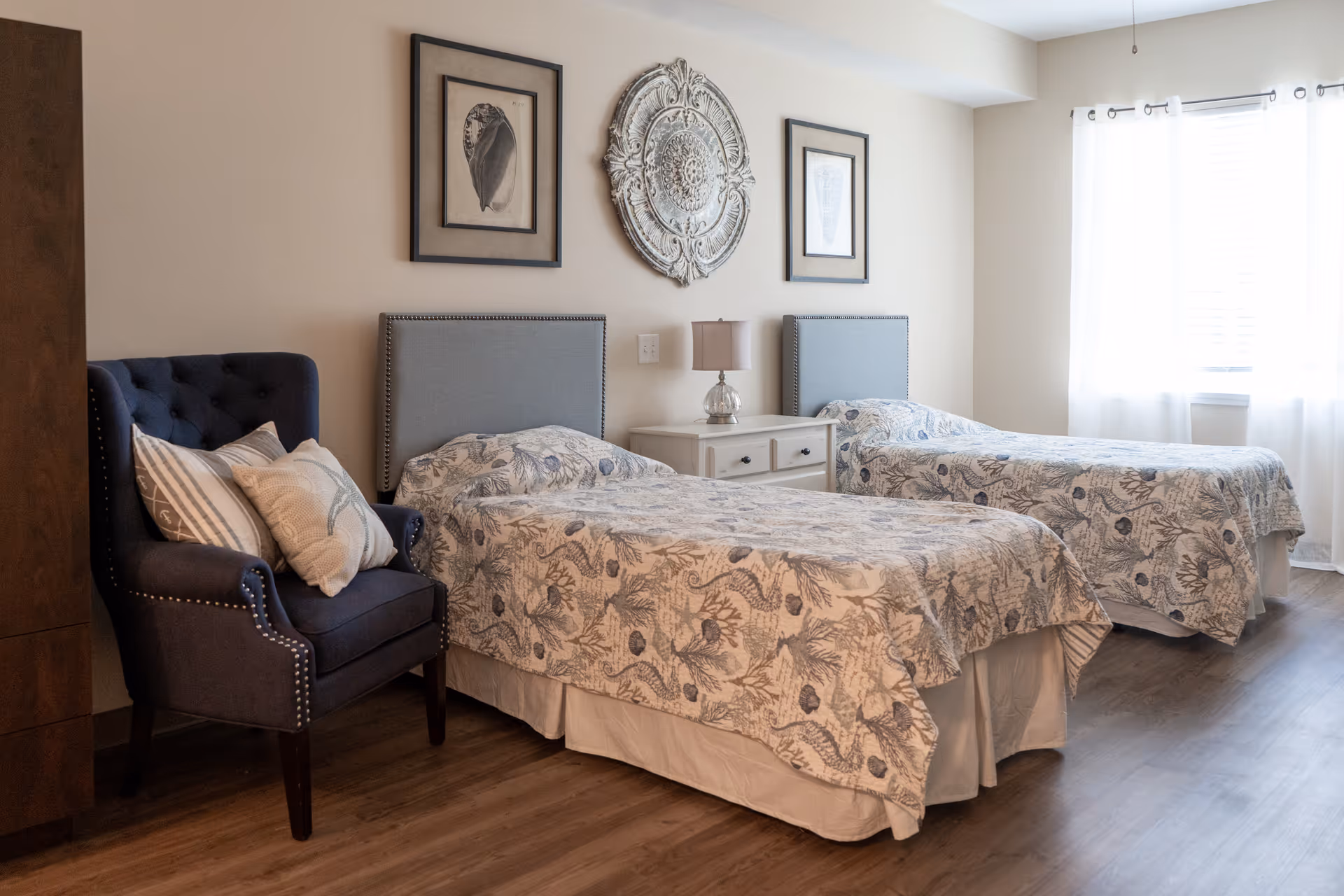 A bedroom with two twin beds featuring light blue patterned bedspreads. Between the beds is a white nightstand with a small lamp. On the left side of the room is a dark blue armchair with three decorative pillows. The walls are decorated with two framed shell prints and a large ornate circular wall decoration. A window with white curtains allows natural light into the room.