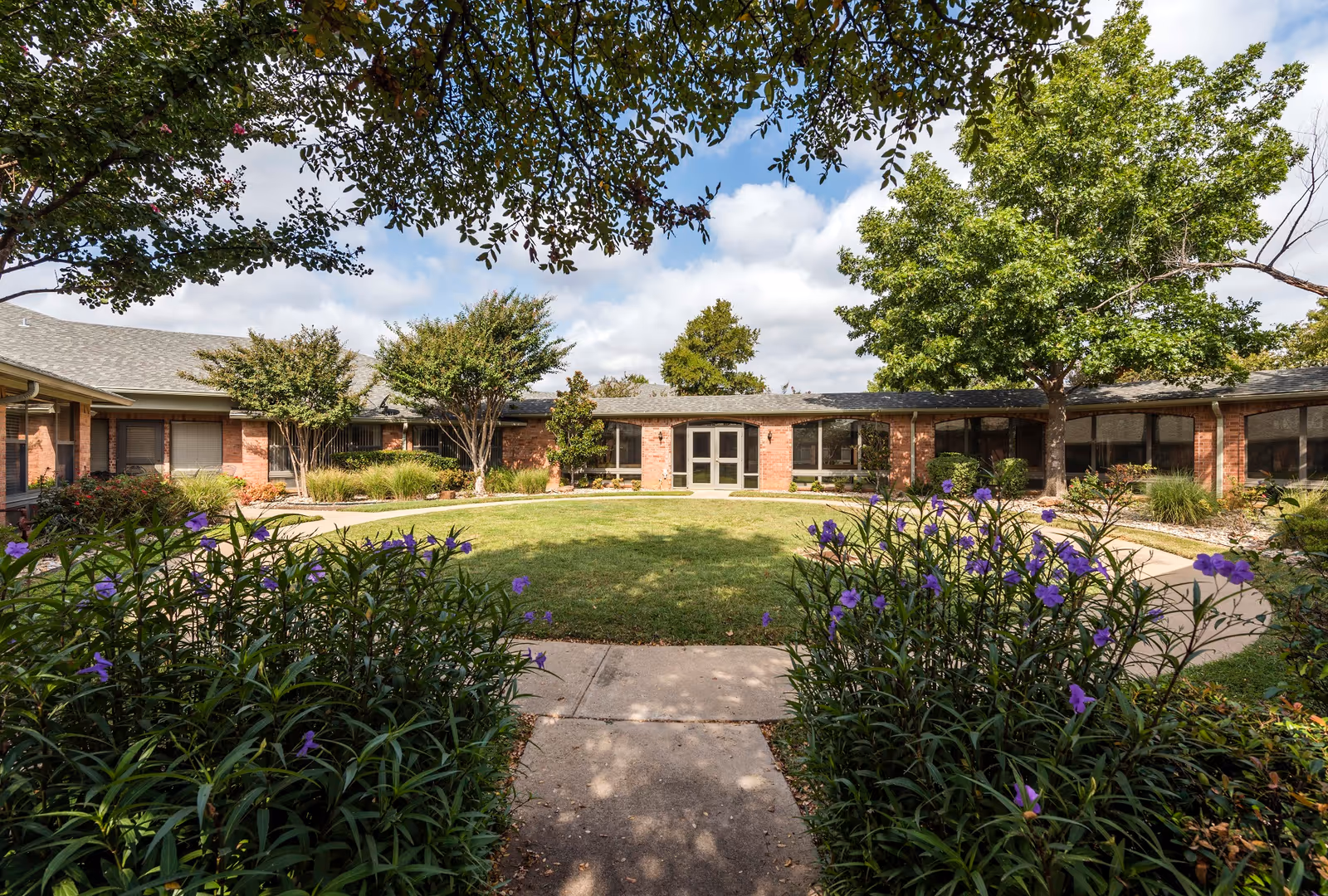 A landscaped courtyard with a circular lawn and purple flowers in front of a low brick senior living facility building.
