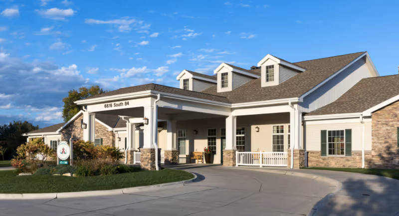 Exterior front view of a single-story senior living facility building with a covered entrance, beige siding, stone accents, and a driveway. The sky is blue with some clouds, and there is a sign on the lawn near the entrance.