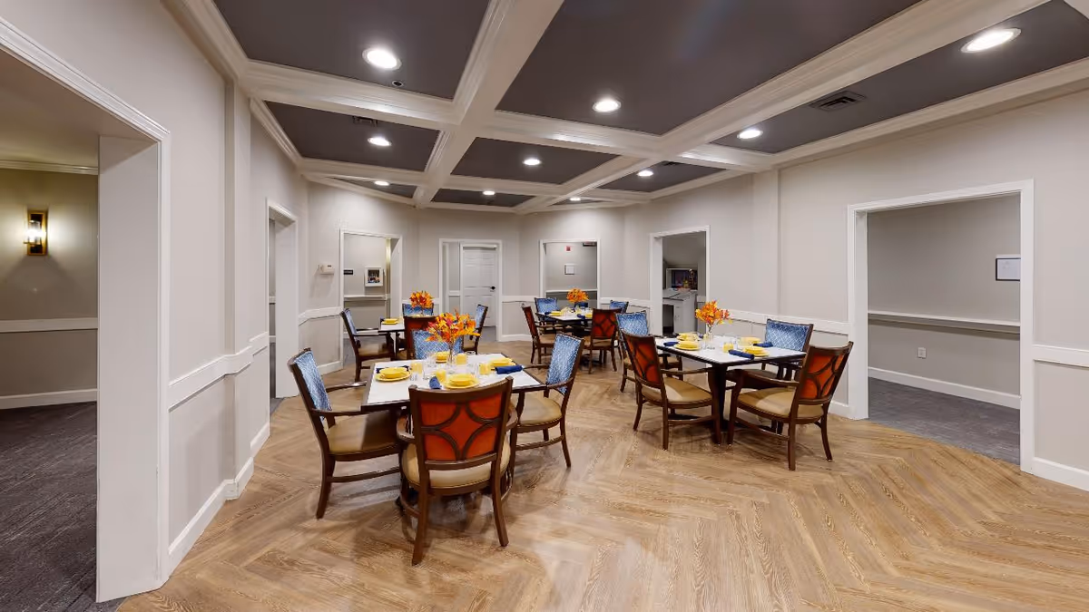 A dining room with several square tables set with yellow plates, blue napkins, glasses, and small flower arrangements. The room has a coffered ceiling with recessed lighting, light-colored walls, and wood-patterned flooring. Multiple doorways lead to adjacent rooms.