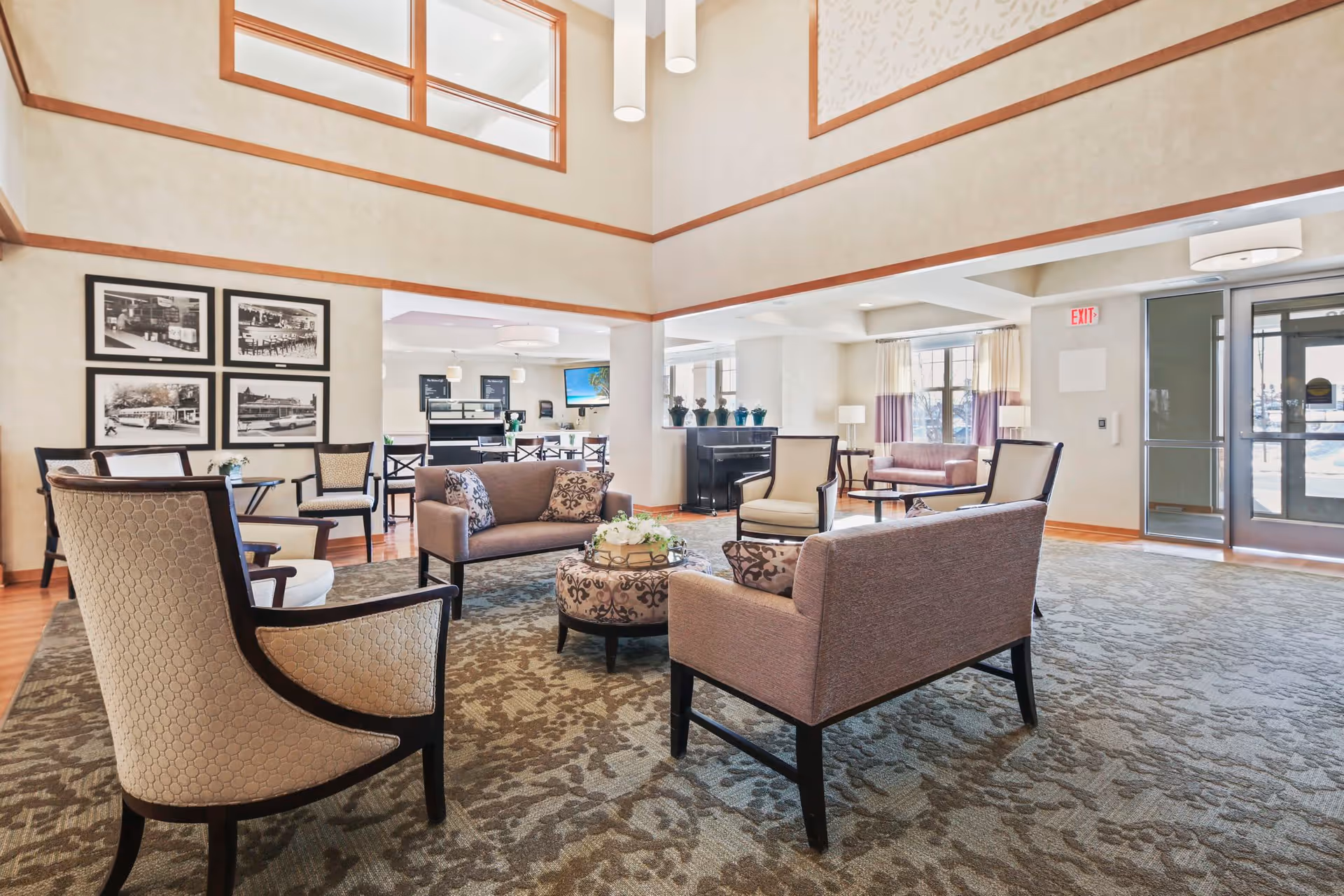 A spacious senior living facility common area with high ceilings and large windows. The room features multiple seating arrangements including beige and brown upholstered chairs and sofas around a central ottoman with a floral arrangement. The walls are decorated with framed black and white photographs. In the background, there is a dining area with tables and chairs, and a piano near the windows. The entrance door with glass panels is visible on the right side.