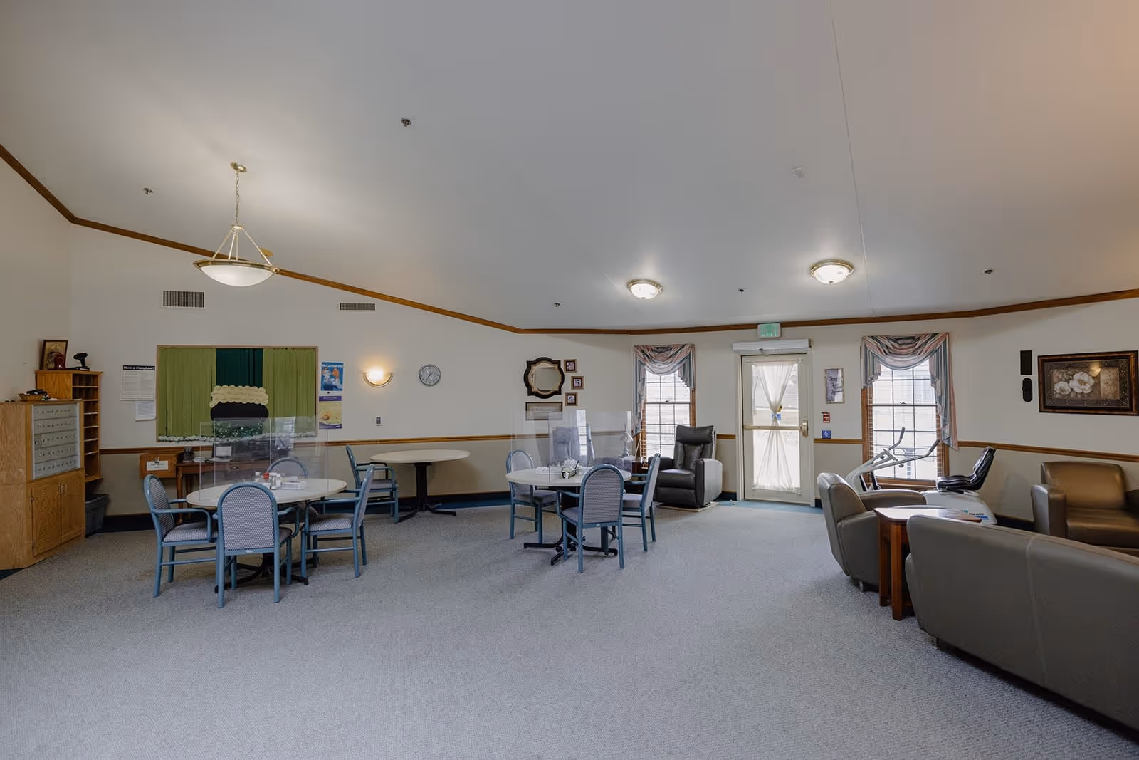 A spacious common area in Redbud Plaza Assisted Living featuring round tables with chairs, some tables equipped with clear protective shields. The room has carpeted flooring, a few armchairs, exercise equipment near windows with draped curtains, and a door leading outside. Walls are decorated with framed pictures, a clock, and a mirror.