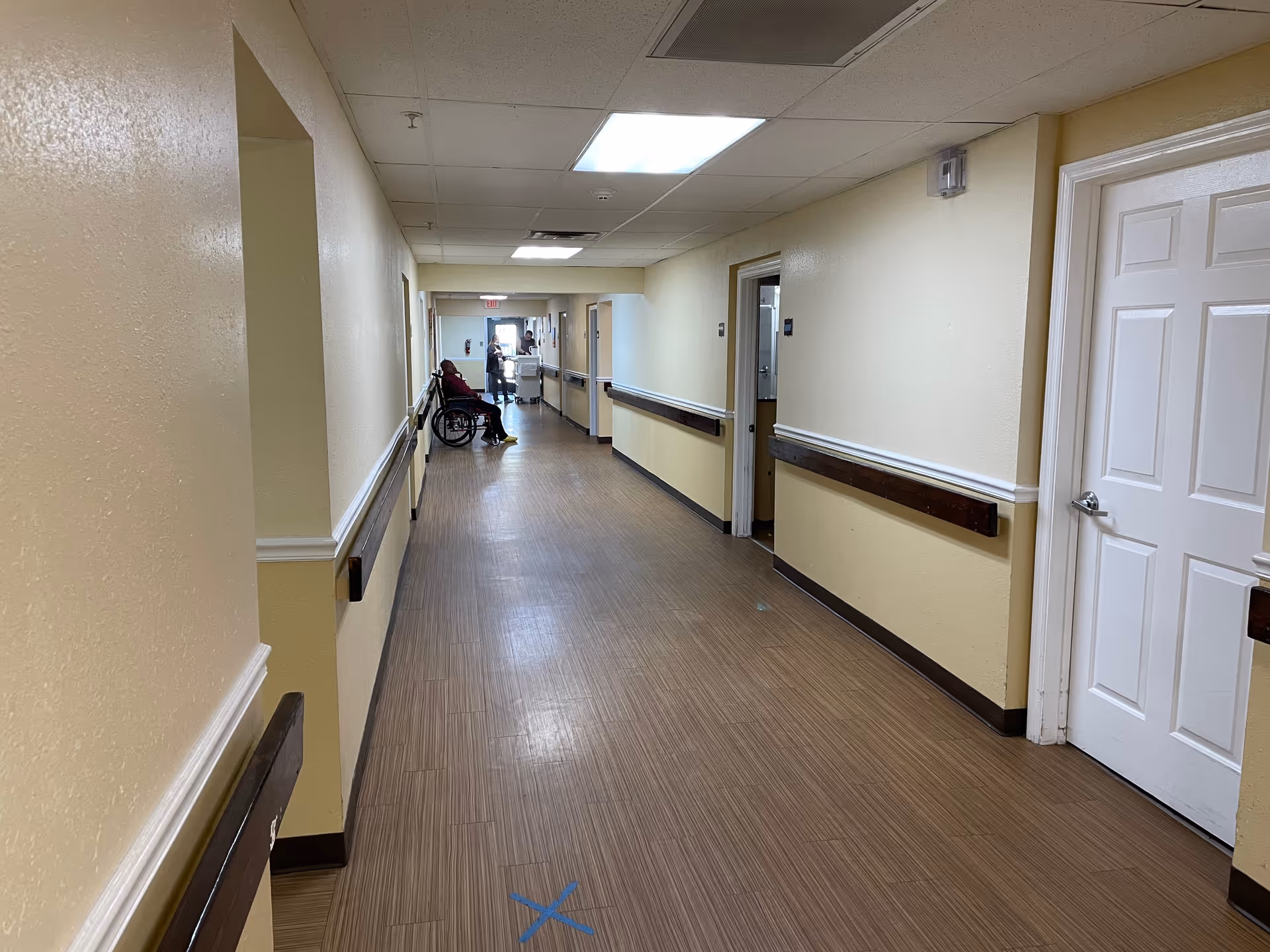 Long interior hallway of an assisted living facility with handrails, doors, and a person in a wheelchair near the far end.