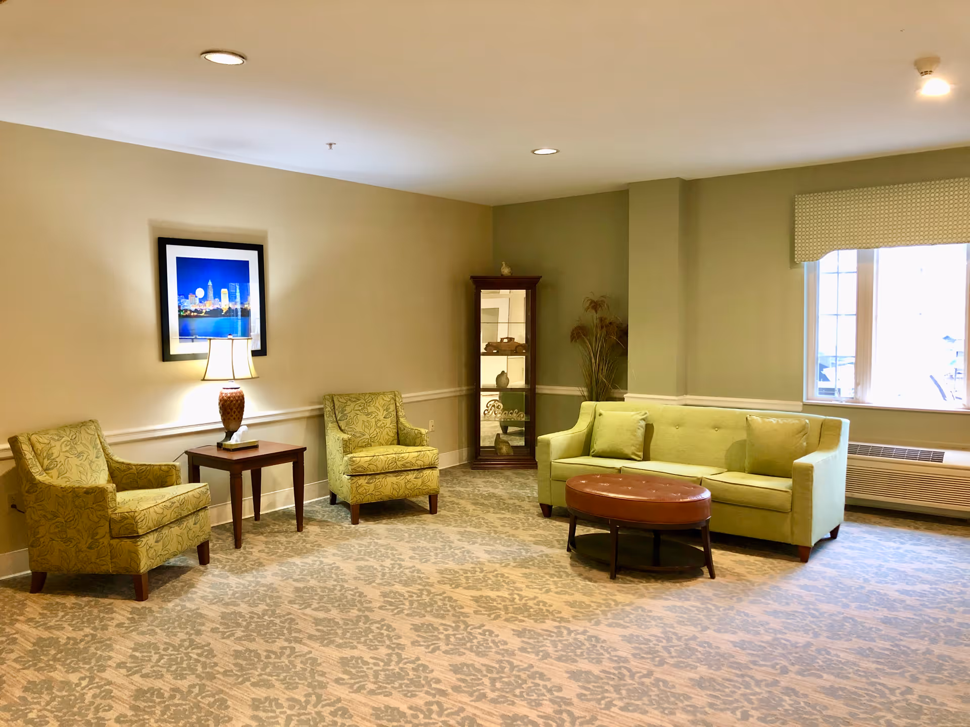 Bright common room with a green sofa, two patterned armchairs, a side table with a lamp, round ottoman and a display cabinet.