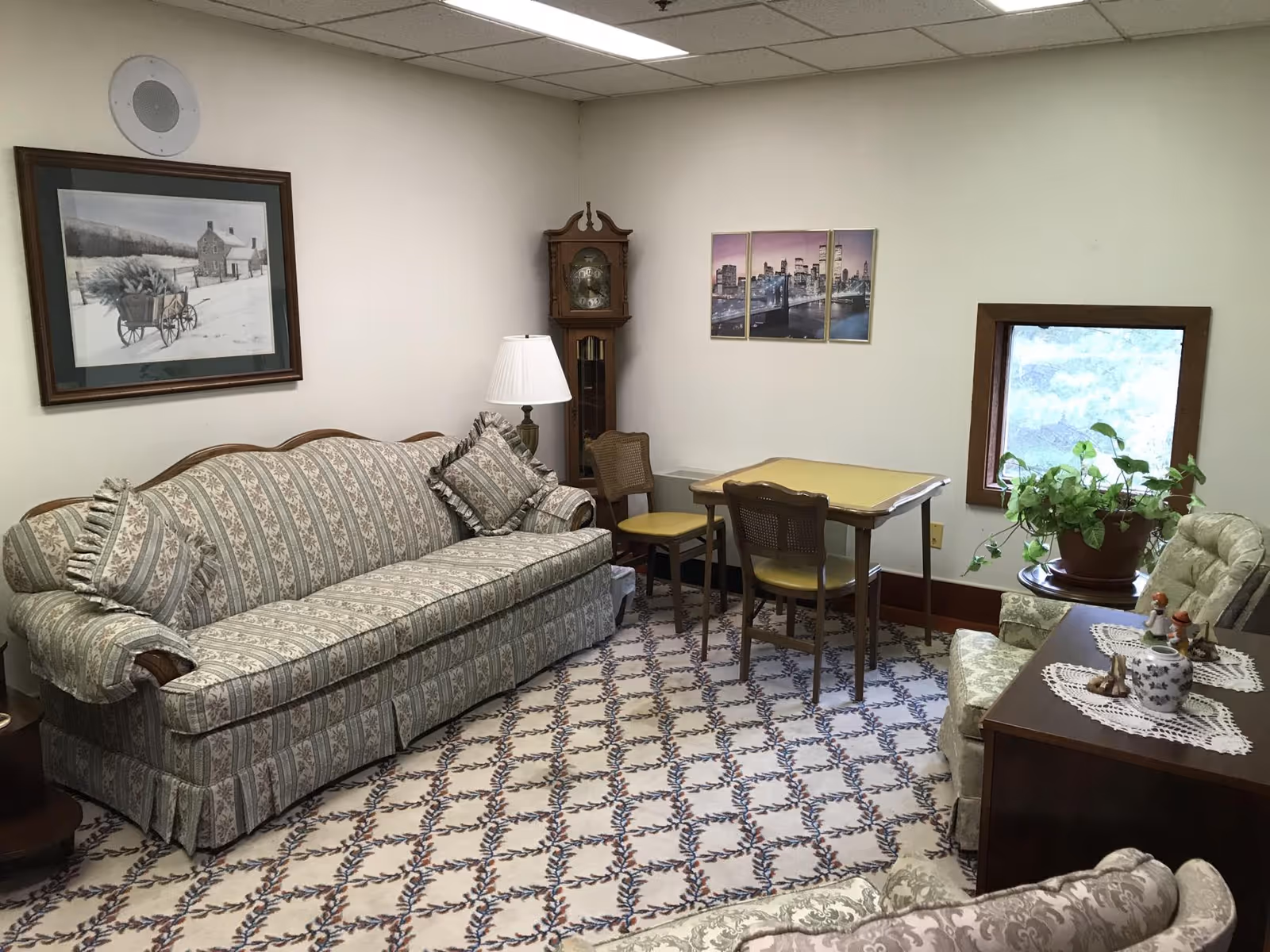 A cozy living room with a patterned sofa and matching cushions, a wooden grandfather clock in the corner, a small table with two chairs, a potted plant on a side table, and framed artwork on the walls. The room has a patterned carpet and a small window letting in natural light.