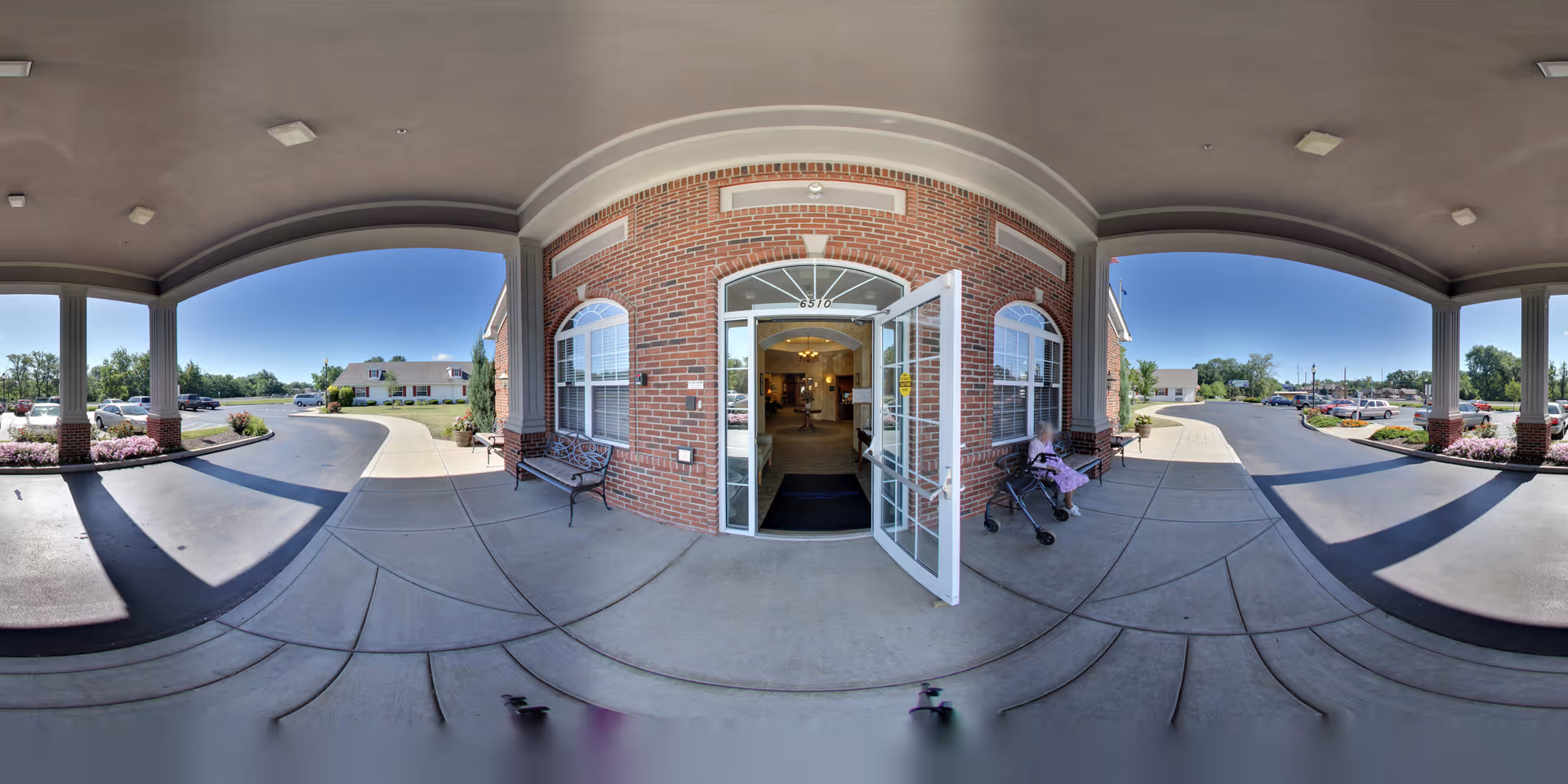 Covered front entrance and drive-up porte-cochère of a brick senior living facility with open glass doors, benches, and a person with a walker.