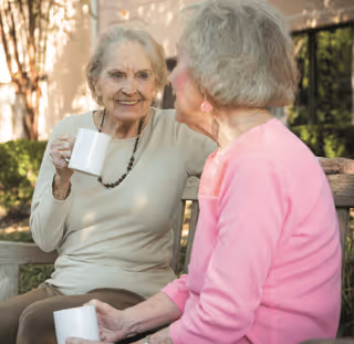 Two elderly women sitting on a wooden bench outdoors, enjoying a conversation while holding white mugs. One woman is wearing a beige top and a necklace, and the other is wearing a pink sweater with matching earrings.