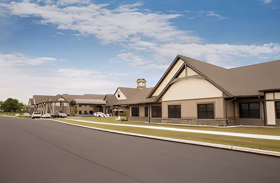 Exterior view of a large, single-story retirement community building with a pitched roof, multiple windows, and a well-maintained lawn under a partly cloudy sky.
