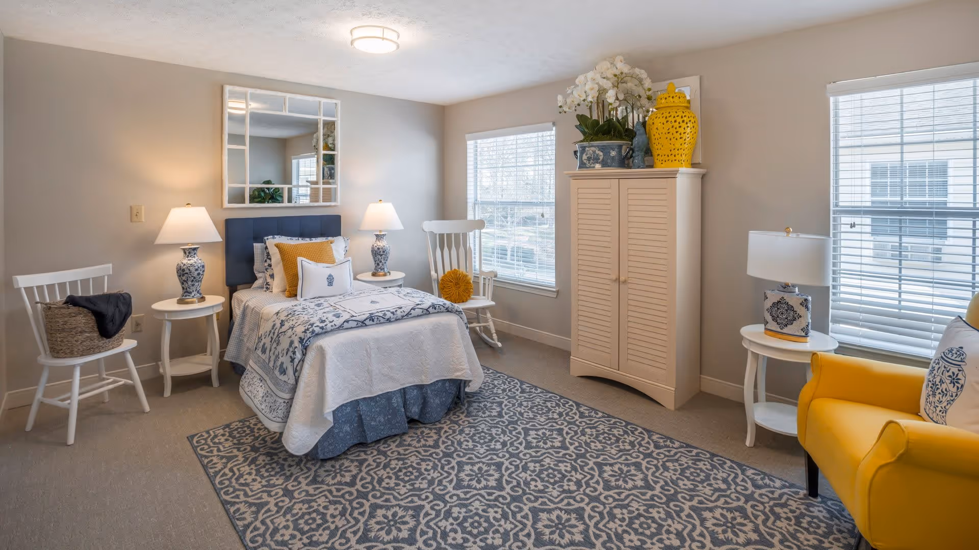 A well-lit bedroom with a single bed dressed in white and blue bedding, flanked by two white side tables each with a blue and white lamp. There are two white chairs, one with a basket and blanket, and the other with a yellow cushion. A large patterned rug covers the floor. A cream-colored cabinet with decorative items on top is positioned between two windows with blinds. A yellow armchair with a decorative pillow is near the right window.