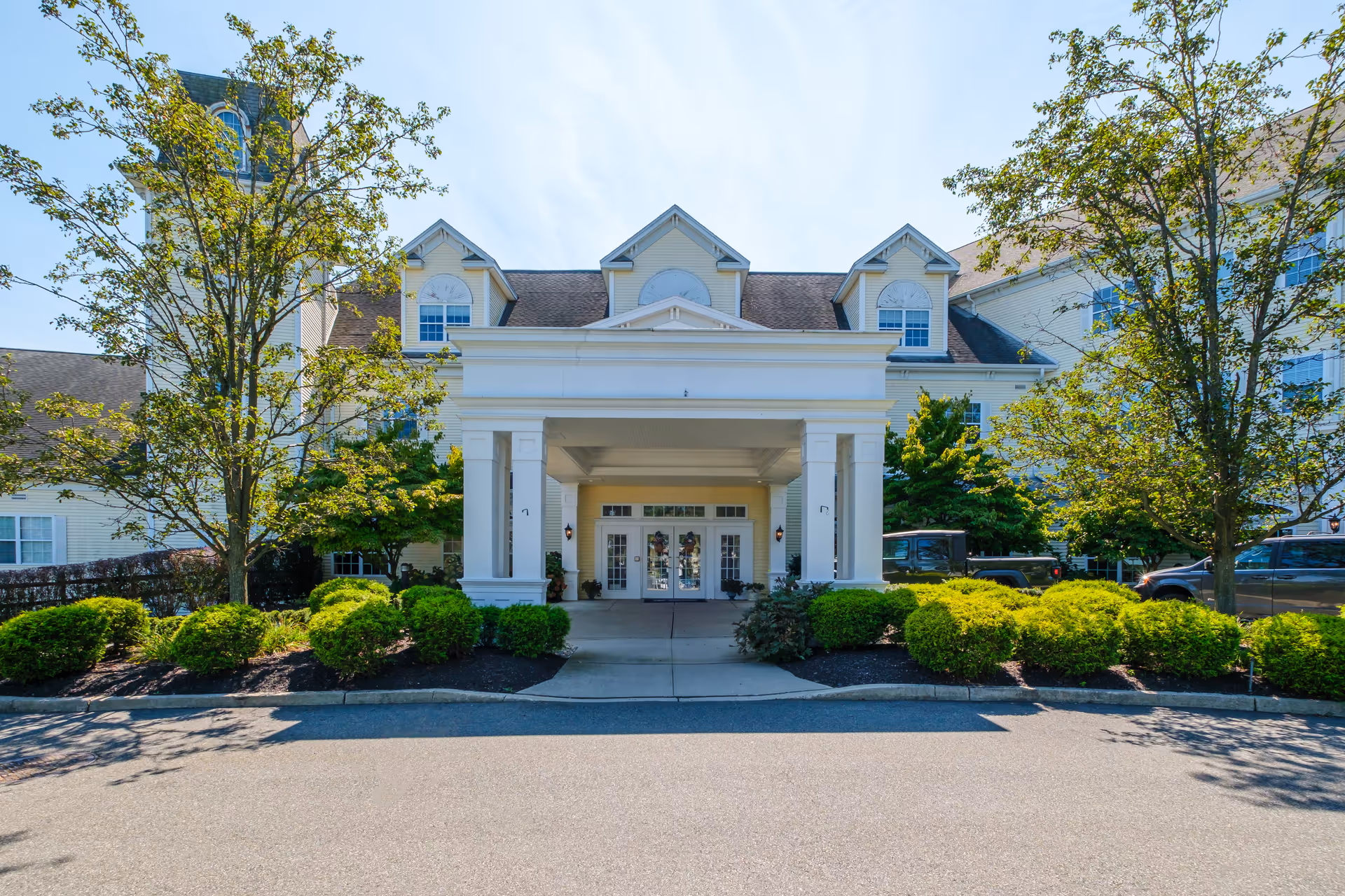 Front entrance of a white multi-story building with a columned porte-cochere, driveway, shrubs, and trees.