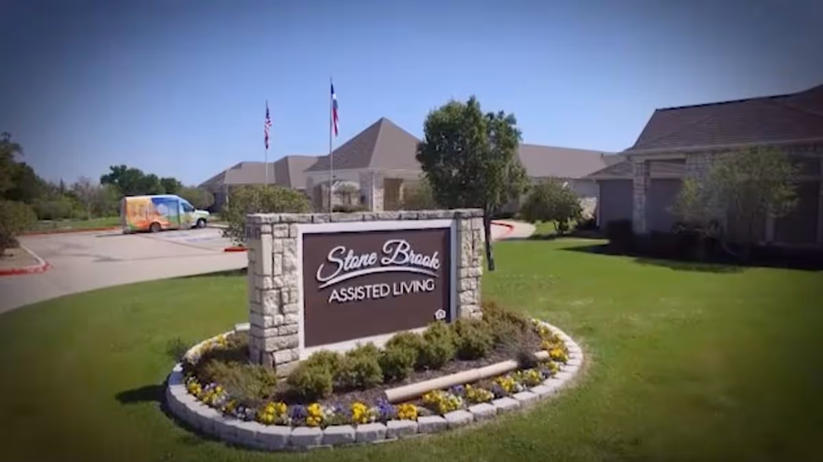 Outdoor view of Stone Brook Assisted Living facility sign surrounded by a flower bed with yellow and purple flowers, green grass, and trees. The building and parking lot with a colorful van are visible in the background under a clear blue sky.