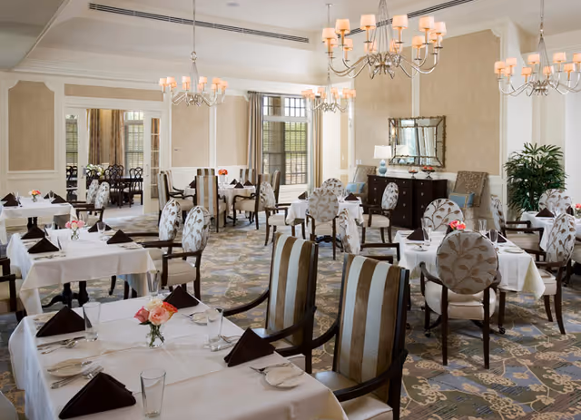 Elegant dining room with multiple tables covered in white tablecloths, each set with plates, glasses, and folded dark napkins. The room features patterned upholstered chairs, chandeliers hanging from the ceiling, large windows with blinds, a decorative mirror on the wall, and a sideboard with lamps and decor items.