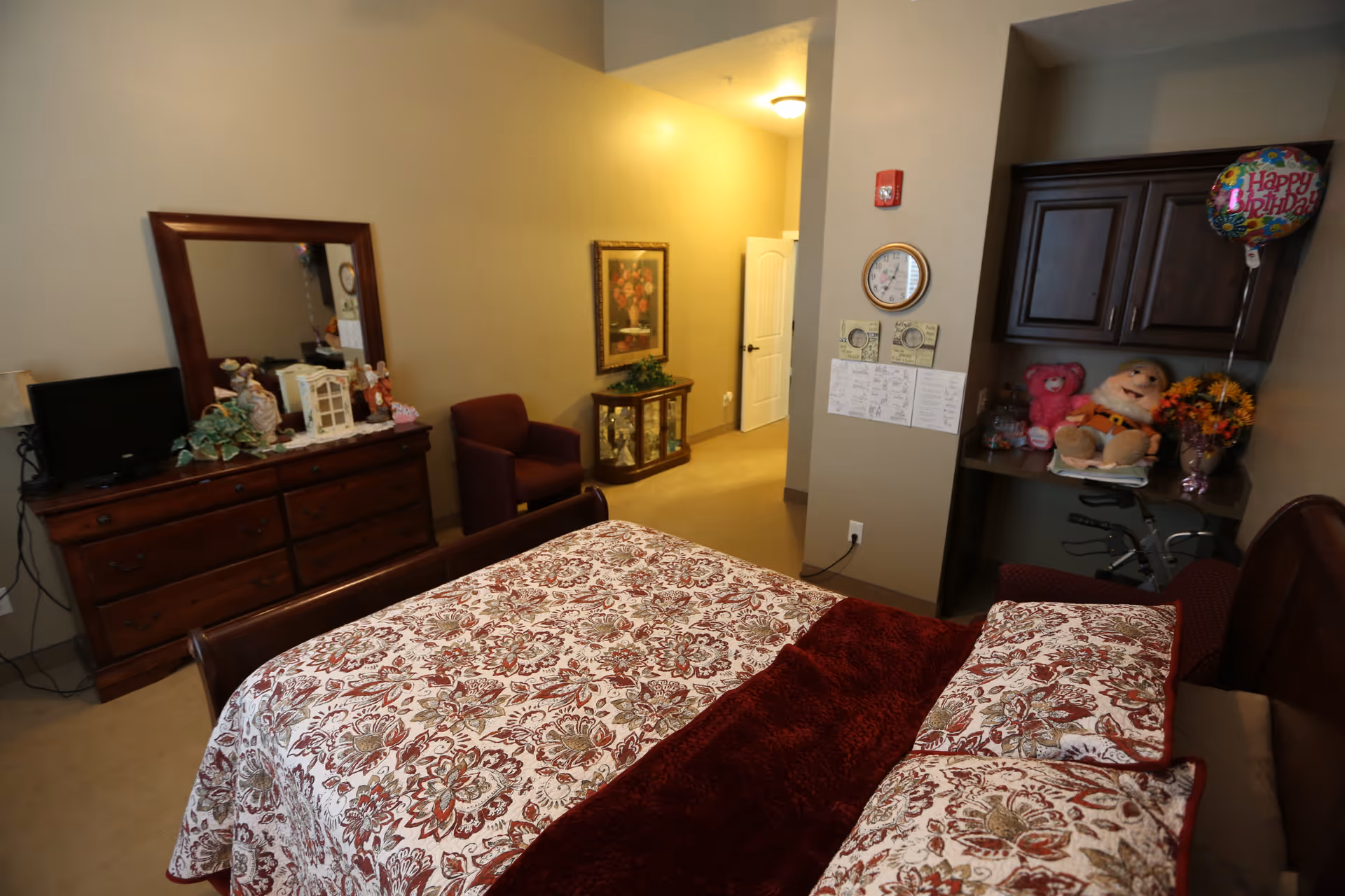 A cozy bedroom in a senior living facility featuring a bed with floral patterned bedding and a burgundy throw blanket. There is a wooden dresser with a mirror and decorative items, a small TV, a burgundy armchair, and a corner desk area with cabinets, stuffed toys, a Happy Birthday balloon, and a flower vase. The room has beige walls and carpeted floor, with a hallway leading to a white door.