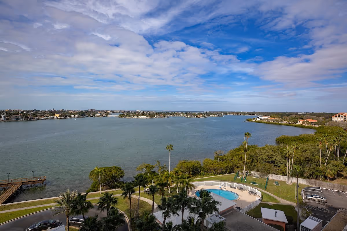 A scenic view overlooking a large body of water with a partly cloudy blue sky. In the foreground, there is a swimming pool surrounded by palm trees, a parking lot with a few cars, and a wooden dock extending into the water. Residential buildings and greenery line the opposite shore.