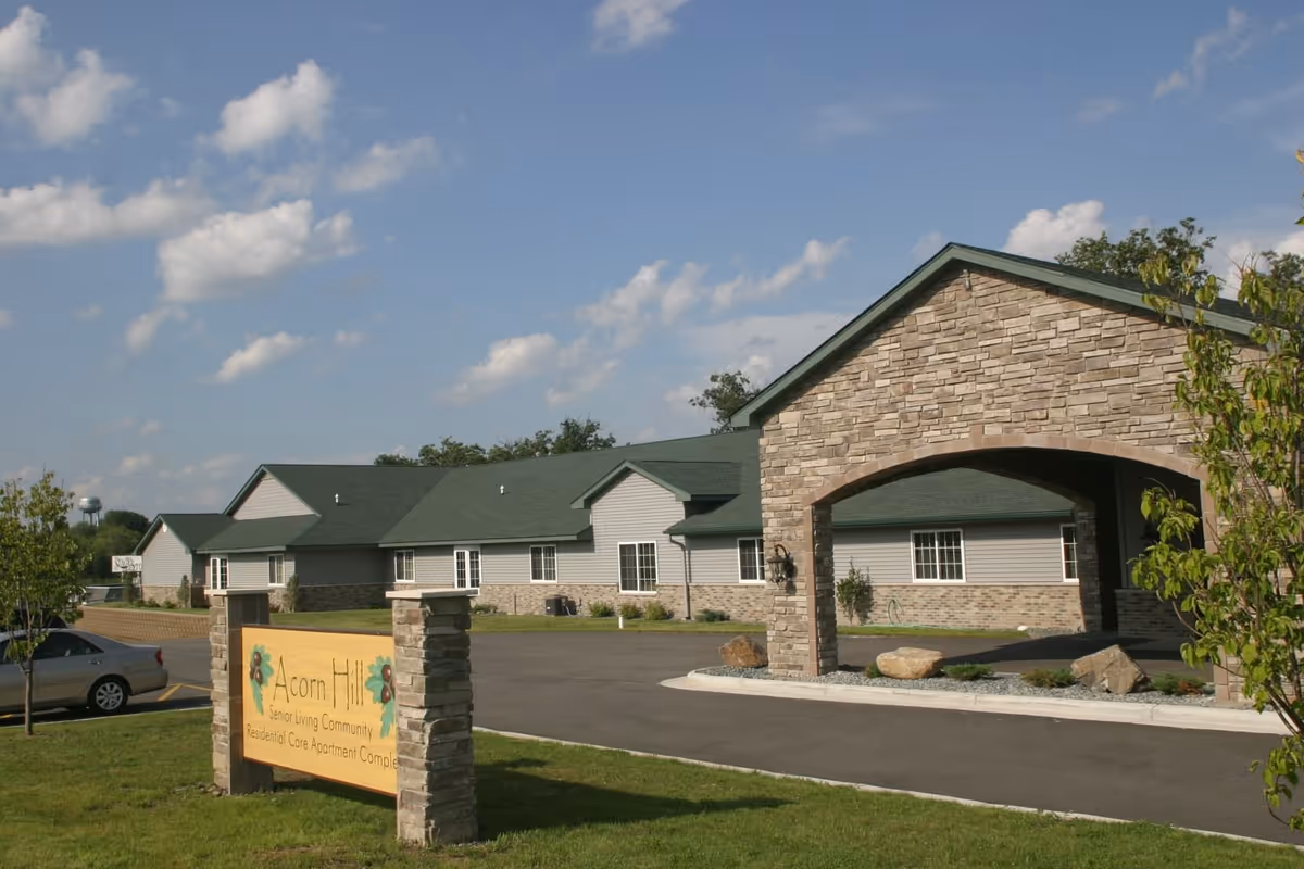 Exterior view of Acorn Hill Apartments senior living community building with stone and siding facade, green roof, and a covered entrance. A sign in front reads 'Acorn Hill Senior Living Community Residential Care Apartment Complex'. There is a car parked on the left side and a blue sky with scattered clouds above.