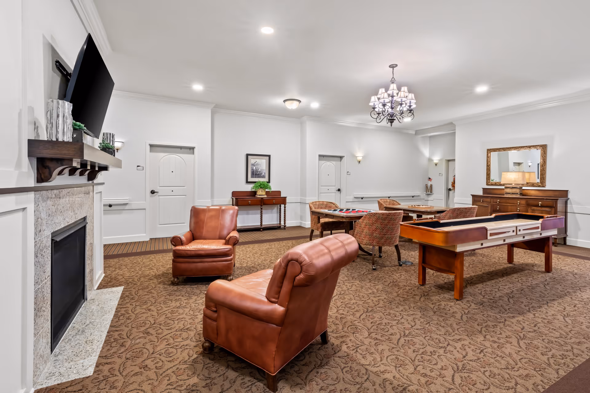 A spacious, well-lit common room with two brown leather armchairs facing a fireplace with a mounted TV above it. The room features a patterned carpet, a shuffleboard table, a table with four upholstered chairs, a wooden sideboard with a lamp and mirror, and white walls with crown molding and recessed lighting.