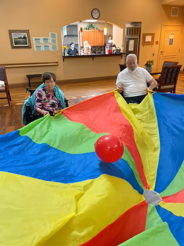 Two elderly residents sit holding a colorful parachute with a red ball in a communal activity room.