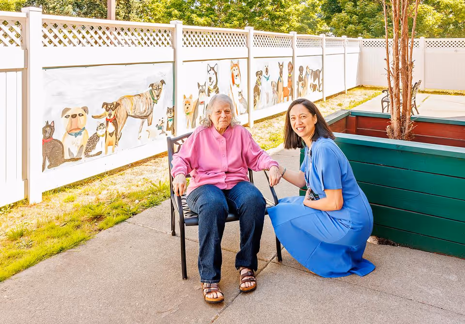 An elderly woman in a pink jacket sitting on a chair outdoors next to a woman in a blue dress who is kneeling and holding her hand. They are in a fenced area with a mural of various dogs painted on the white fence behind them.