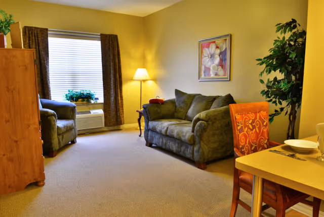 A cozy living room with a green upholstered loveseat and armchair, a wooden cabinet, a floor lamp, a framed floral painting on the wall, a window with brown curtains, a potted plant, and a dining table with an orange patterned chair and place setting.