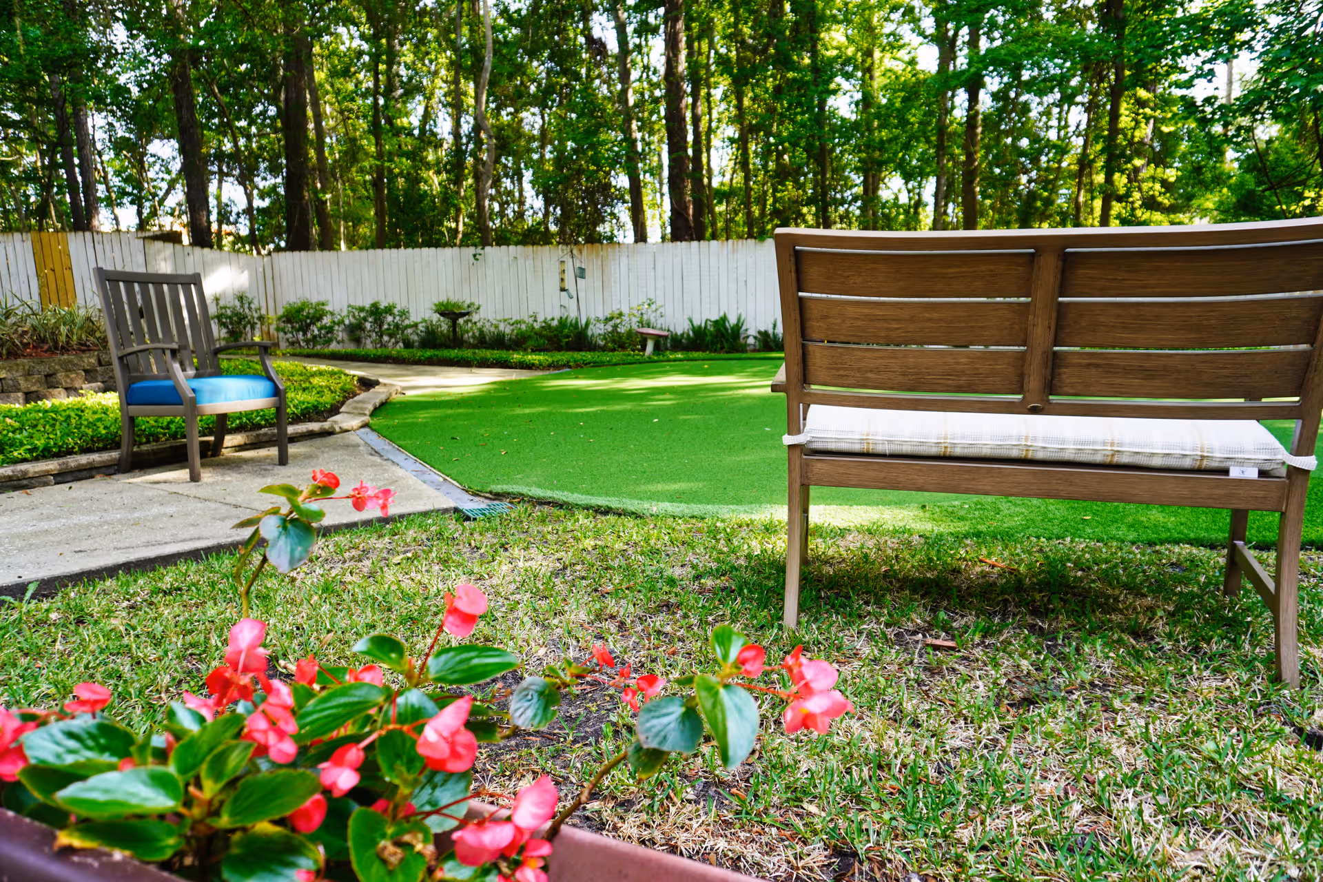 A peaceful outdoor garden area with a wooden bench featuring a cushion and a wooden chair with a blue cushion. There are vibrant pink flowers in the foreground, a green artificial turf area, a concrete pathway, and a white wooden fence with trees in the background.