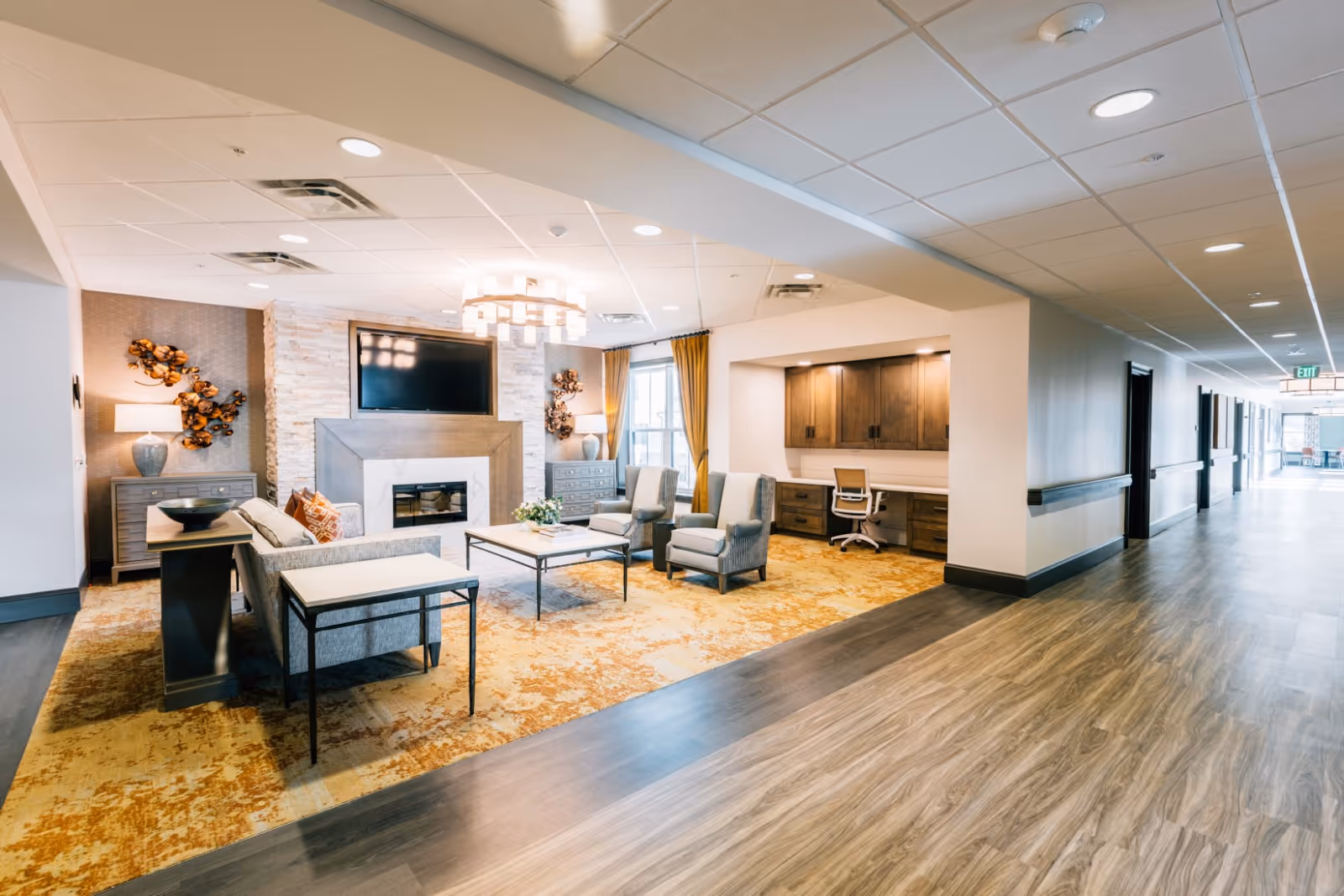 Bright communal lounge with seating, a fireplace and wall-mounted TV opening onto a wide hallway.
