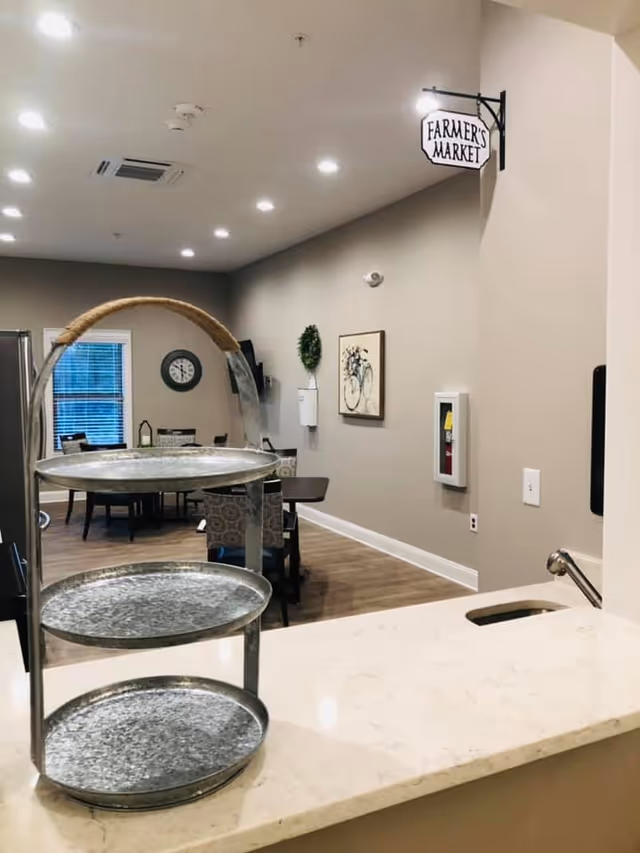 Interior view of a senior living facility dining area with a three-tiered metal tray on a marble countertop in the foreground. The room has beige walls, wooden flooring, and recessed ceiling lights. A small sign reading 'FARMER'S MARKET' hangs on the wall. In the background, there are tables and chairs, a wall clock, a framed bicycle artwork, and a small green wreath.