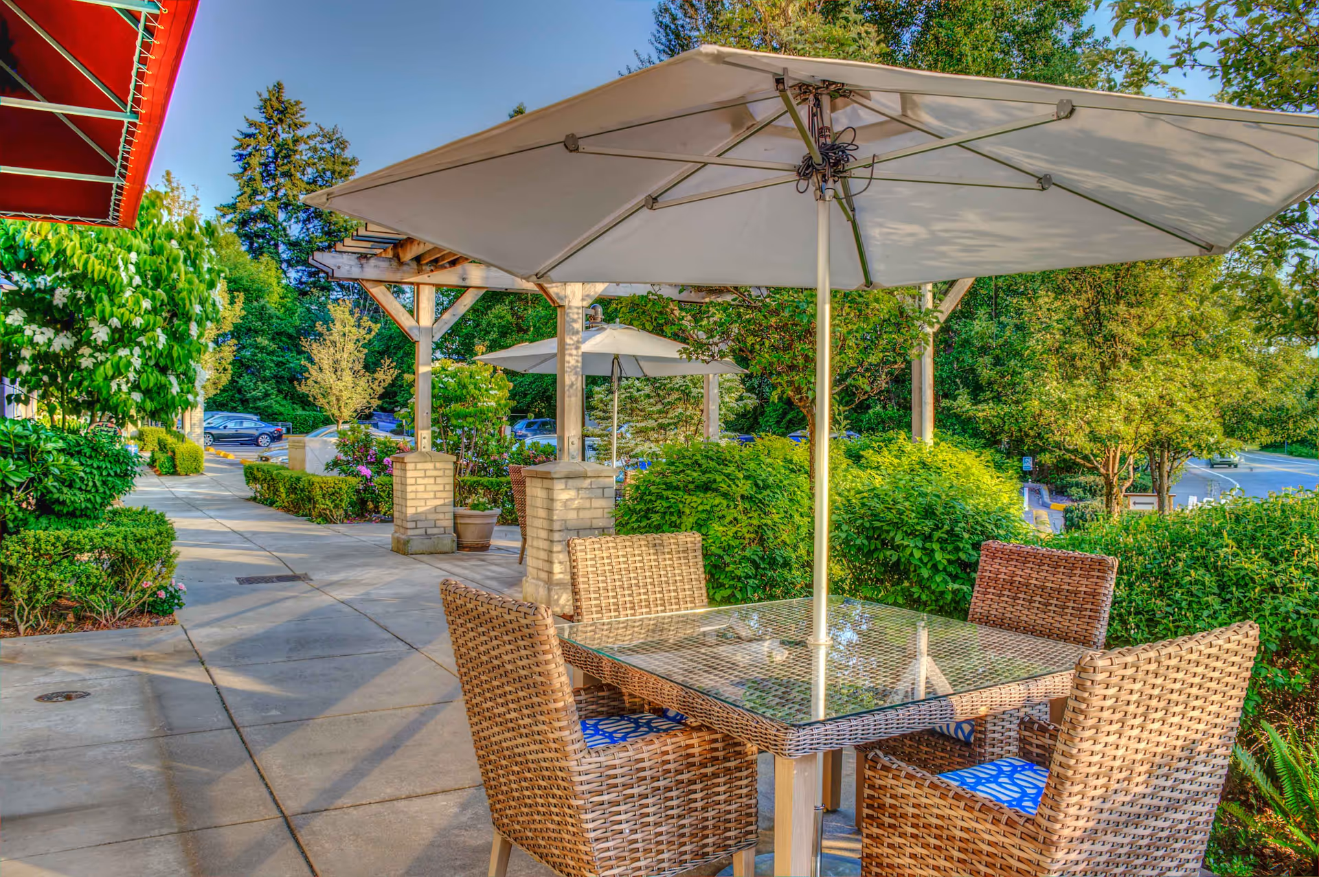 Outdoor patio with a glass-top table, four wicker chairs and a large umbrella beside a walkway and landscaped greenery.