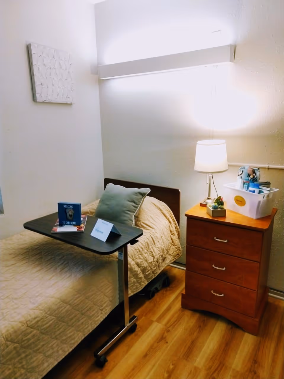A small, cozy bedroom with a single bed covered in a beige quilt and a green pillow. A dark wooden overbed table is positioned over the bed, holding a small welcome card and another card. Next to the bed is a wooden nightstand with three drawers, a lamp, a small plant, and a white plastic bin containing various items. The room has light-colored walls and wood flooring.