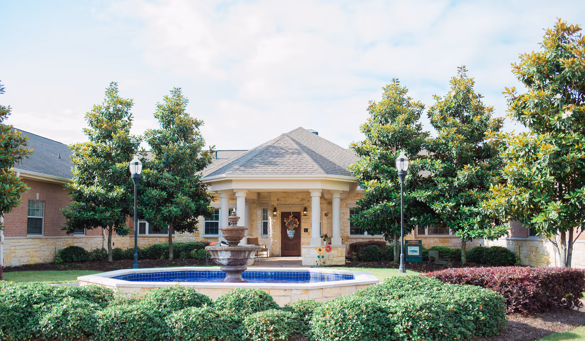 Front entrance of a single-story senior care building with a central fountain, columns, and landscaped trees and shrubs.