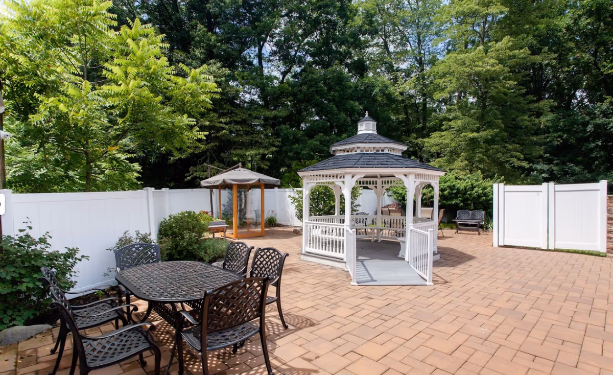 Outdoor patio area with a white gazebo in the center, surrounded by trees and greenery. There is a black metal table with six matching chairs on the left side and a small wooden structure with a canopy in the background. The area is paved with light brown bricks and enclosed by a white fence.