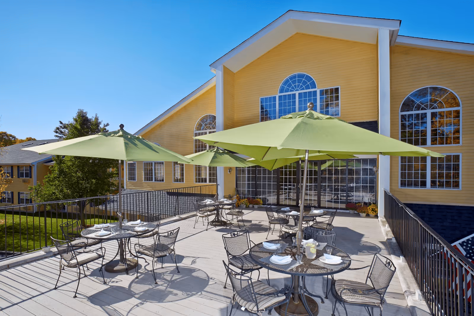 Outdoor patio area with several round metal tables and chairs, each table shaded by large green umbrellas. The patio is attached to a yellow building with large arched windows and a clear blue sky overhead.