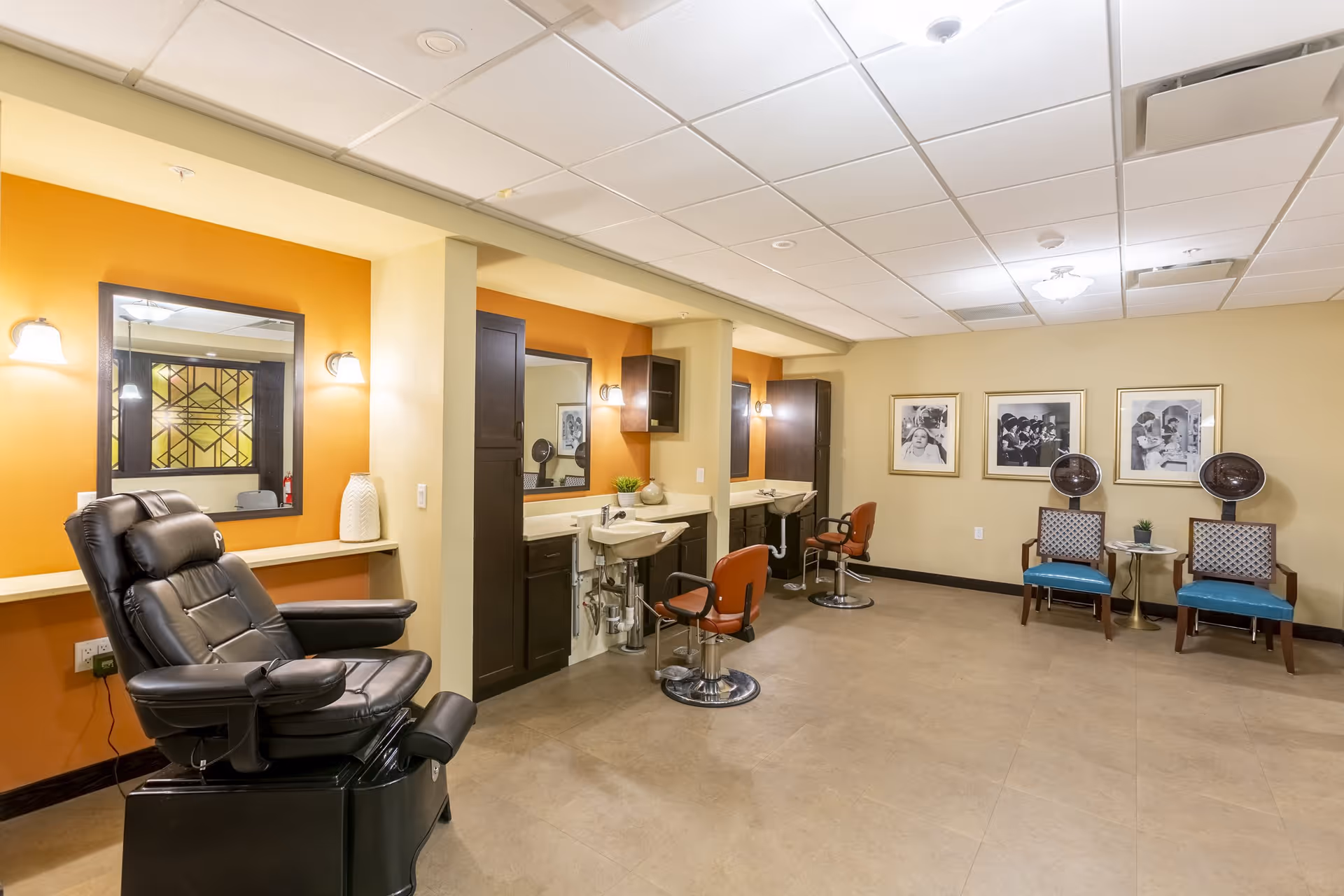 Interior view of a salon area in a senior living facility with a black reclining chair, two salon chairs in front of sinks and mirrors, two blue chairs with hair dryers, and framed black and white photos on the wall.