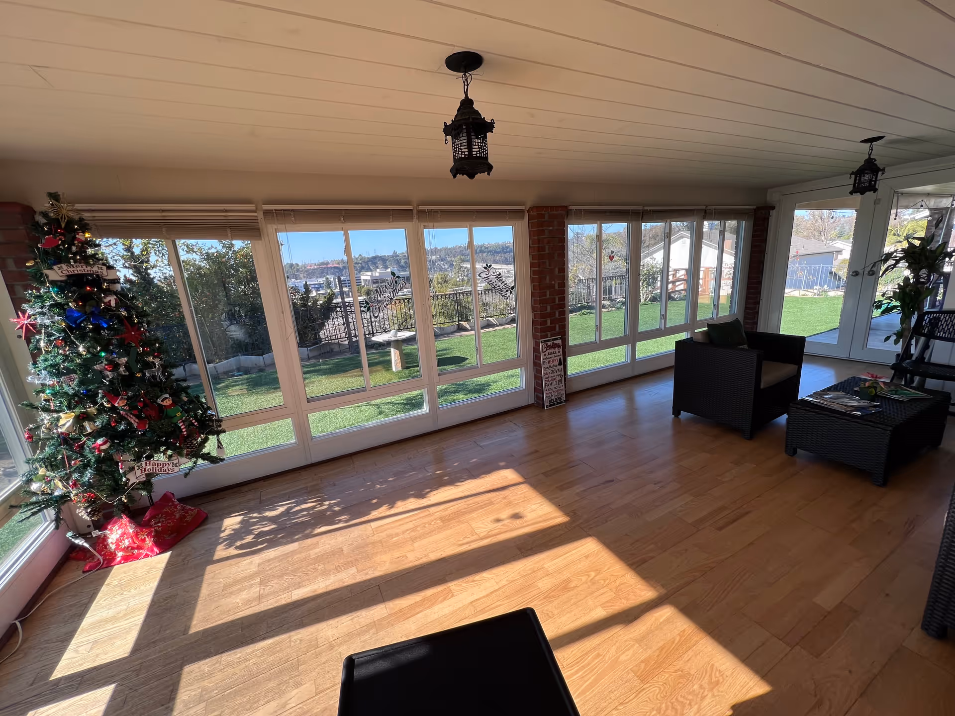 Bright sunroom with large windows overlooking a lawn, wicker seating, and a decorated Christmas tree.