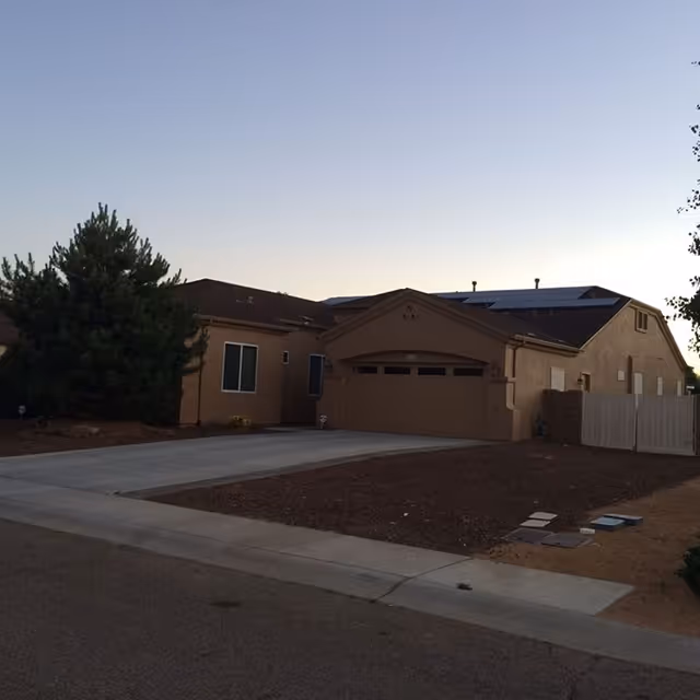 Exterior view of a single-story residential building with a two-car garage, a concrete driveway, and a tree on the left side. The sky is clear and it appears to be either early morning or late evening.