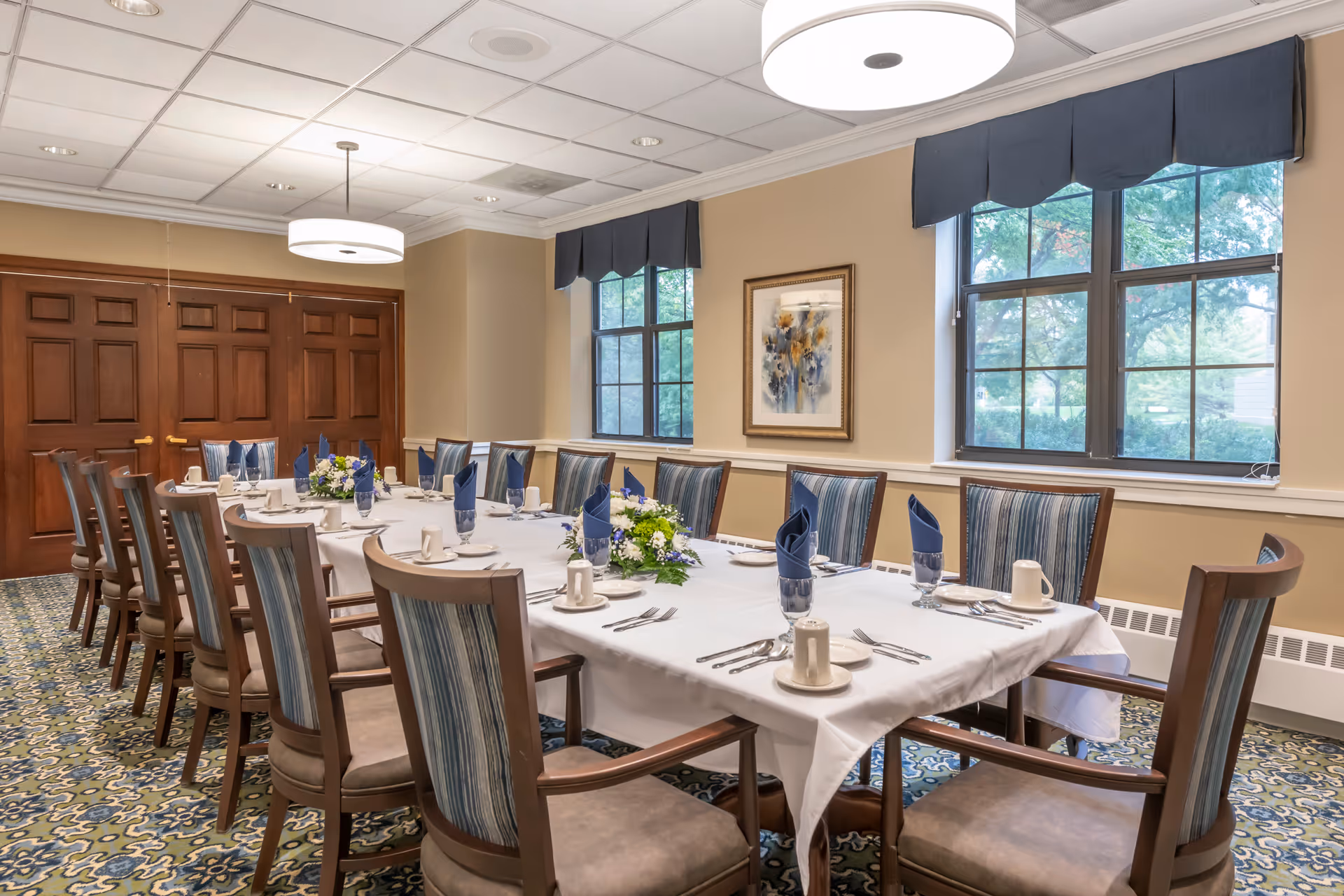 A formal dining room with a long rectangular table covered with a white tablecloth. The table is set with plates, cups, silverware, blue napkins, and floral centerpieces. The room has patterned carpet, beige walls, large windows with blue valances, and wooden double doors.