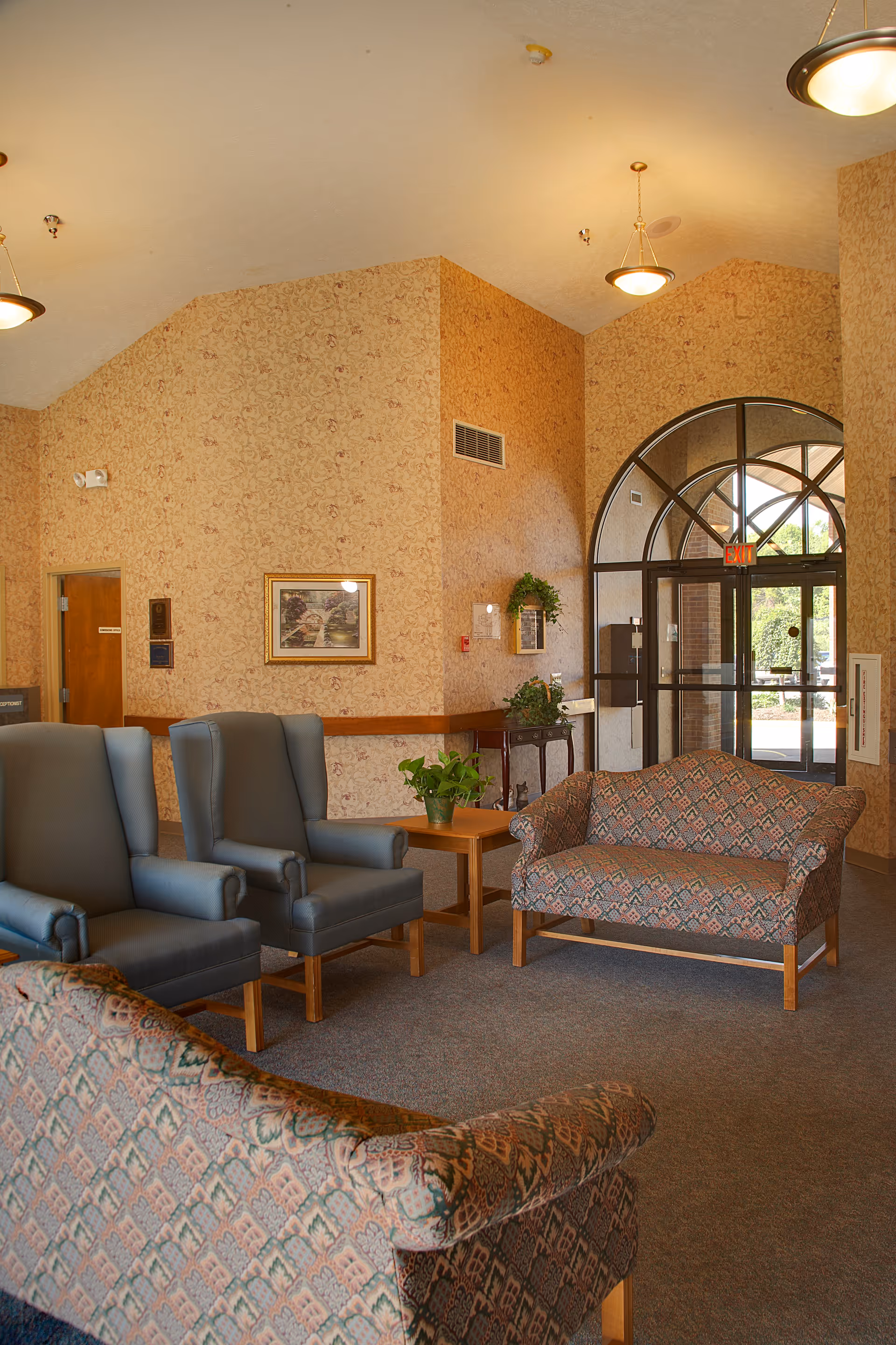 A cozy seating area in a senior living facility with patterned sofas and blue armchairs arranged around a wooden side table with a potted plant. The room has beige floral wallpaper, a framed picture on the wall, and a large arched glass door leading outside. Ceiling lights illuminate the space.