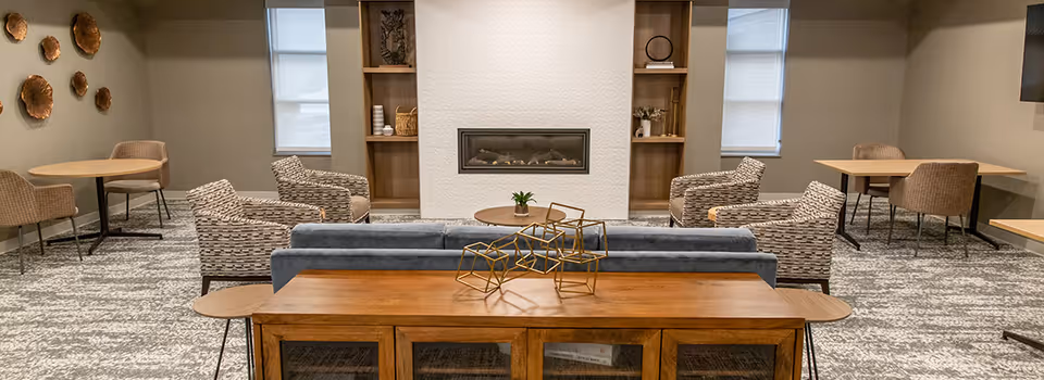 A modern and cozy common area with a gray sofa, four patterned armchairs, two round wooden tables with chairs, a wooden console table with decorative geometric sculptures, built-in shelves, and a wall-mounted fireplace in the center.