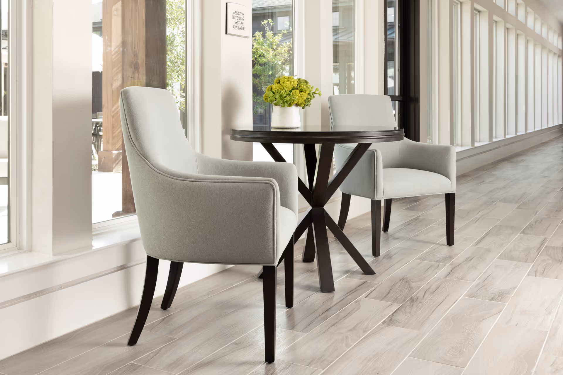 A small round dark wood table with a white vase holding green flowers on top, flanked by two light gray upholstered armchairs with dark wooden legs, set in a bright hallway with large windows and light wood flooring.