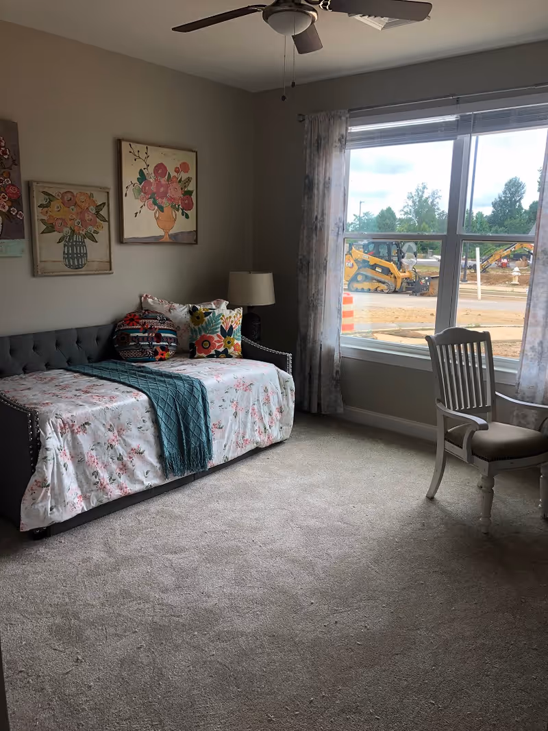 Sunlit bedroom with a daybed dressed in floral bedding and colorful pillows, a lamp and chair by a large window showing construction outside.