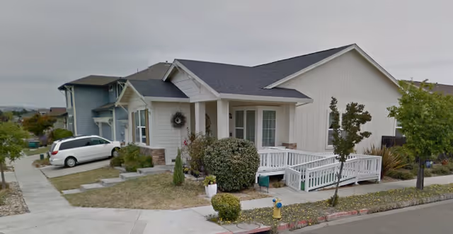 Exterior view of a single-story residential building with a white wooden ramp and porch, surrounded by shrubs and small trees. A white minivan is parked on the driveway next to the house, and another two-story house is visible in the background.