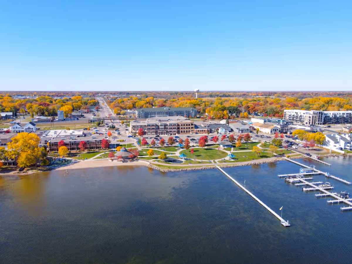 Aerial view of a lakeside town with colorful autumn trees, a park with walking paths and gazebos along the shoreline, residential and commercial buildings, and a marina with docks extending into the water under a clear blue sky.