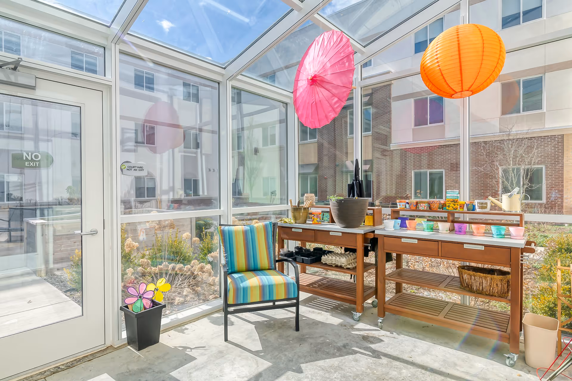 A bright sunroom with glass walls and ceiling showing an outdoor courtyard. Inside, there is a colorful striped chair and two wooden tables on wheels holding various gardening supplies, pots, and small colorful containers. Two decorative paper lanterns, one pink and one orange, hang from the ceiling. Outside the glass, there are plants and a building with multiple windows.
