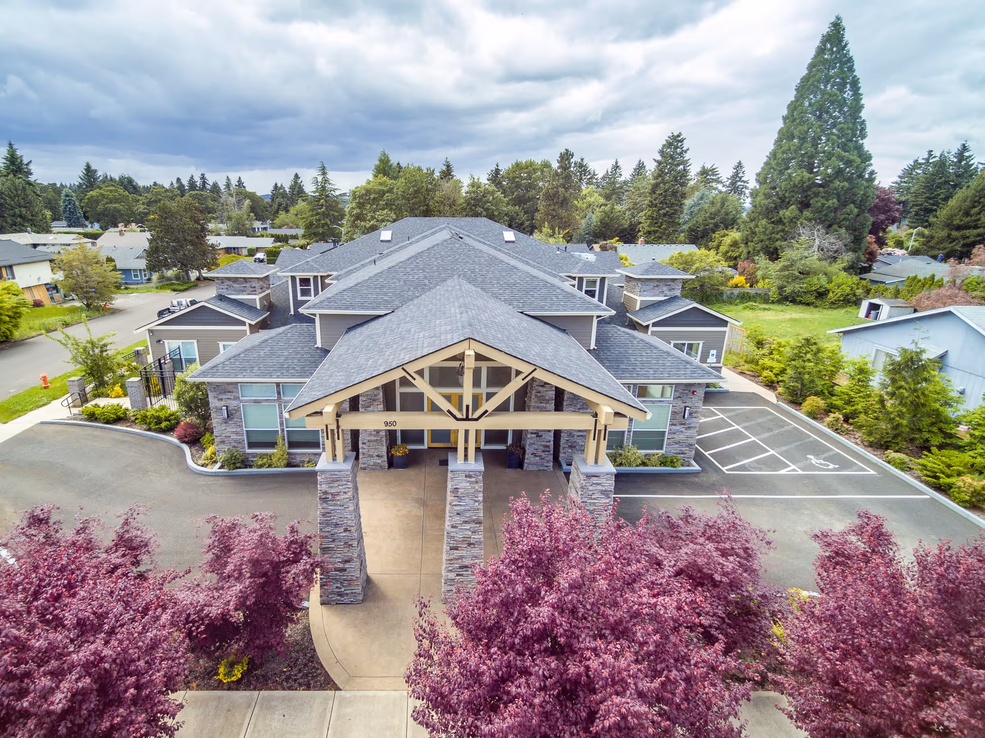 Aerial view of the front entrance of a single-story senior living facility building with a covered driveway supported by stone pillars. The building is surrounded by trees and landscaping, with a parking area including a handicapped parking space visible to the right. The sky is partly cloudy.