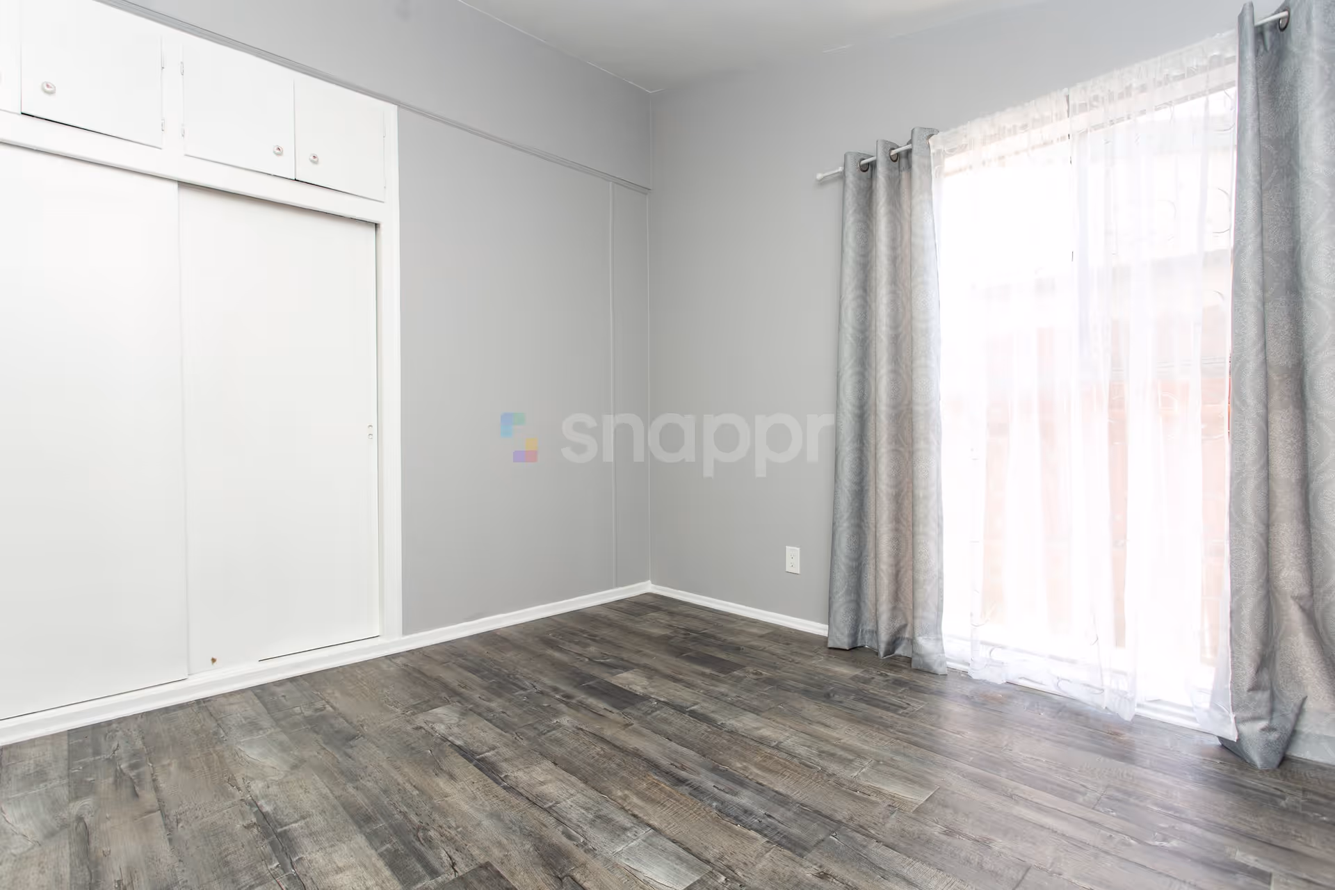 Empty room with gray walls, dark wood flooring, a large window covered with sheer white curtains and gray drapes, and a white built-in closet with sliding doors.