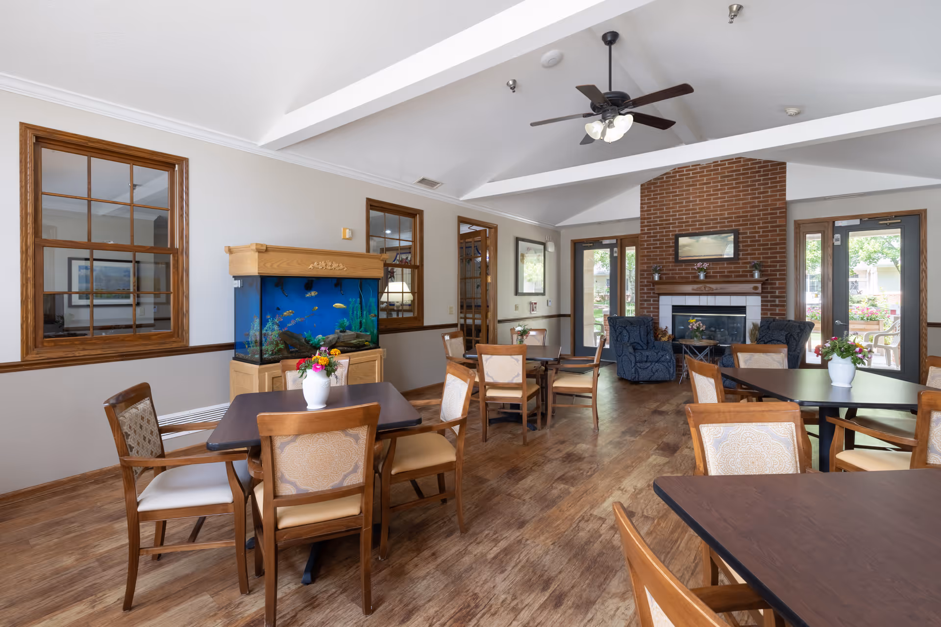 A bright and spacious common area with wooden floors, several tables and chairs arranged for seating, a large fish tank against the wall, a brick fireplace with two blue armchairs nearby, and large windows and glass doors letting in natural light.