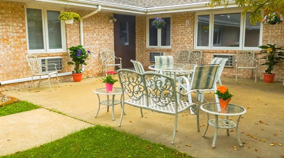 Outdoor patio area with metal chairs and tables, some with cushions, surrounded by brick walls with windows and air conditioning units. Several potted plants and hanging flower baskets decorate the space.