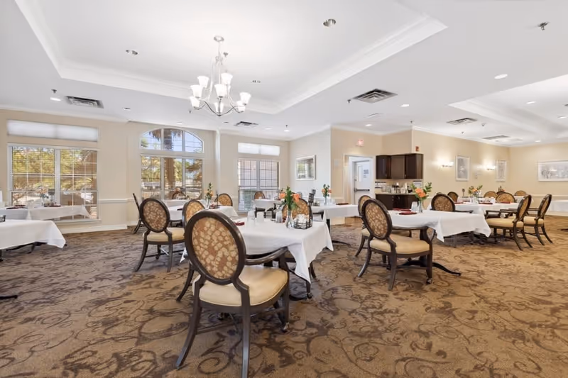 A spacious dining room with multiple tables covered in white tablecloths, each set with chairs featuring patterned upholstery. The room has large windows allowing natural light to fill the space, a chandelier hanging from the ceiling, and a carpeted floor with a decorative pattern. There are small flower arrangements on each table and a counter area with cabinets in the background.